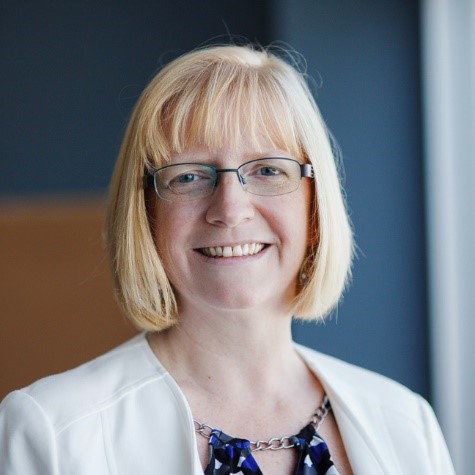 Professional headshot of a smiling woman with shoulder-length blonde hair and glasses, wearing a white blazer and a statement necklace.