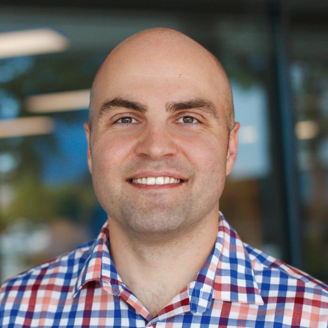 A smiling man with a shaved head wearing a checkered shirt.