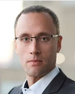 A professional headshot of a man with short hair and glasses, wearing a suit jacket and white shirt.