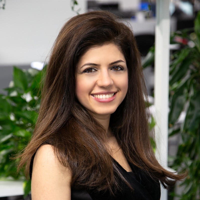 Smiling woman with long brown hair in a black top, photographed indoors with greenery in the background.