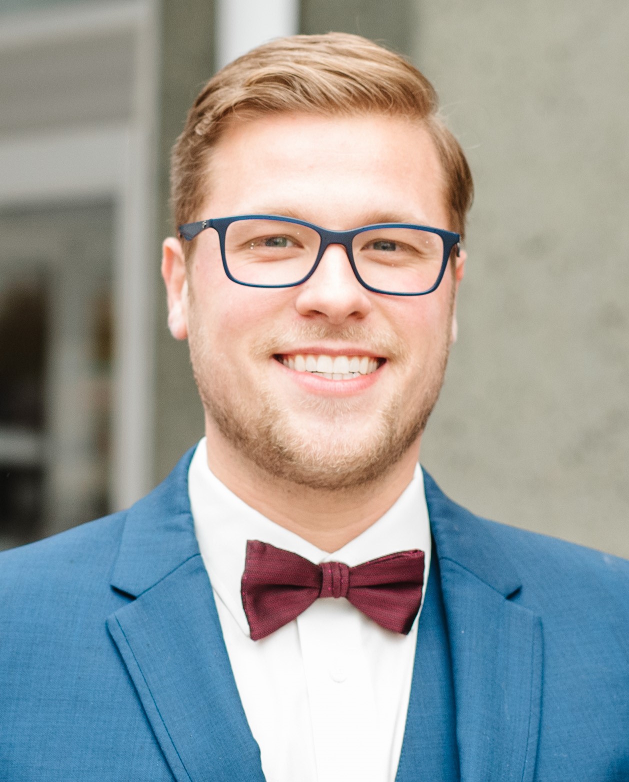 Smiling person wearing blue-framed glasses, a blue suit jacket, and a burgundy bow tie in a close-up portrait.