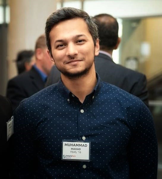 A young man with a beard, wearing a navy blue button-up shirt with white polka dots, stands smiling at a networking event, with a name tag that reads "Muhammad Mahad FEA'S '16."