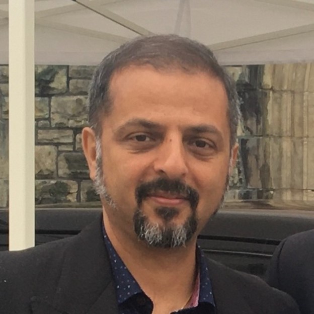 A portrait of a man with a beard, wearing a black suit and a patterned shirt, smiling outdoors.