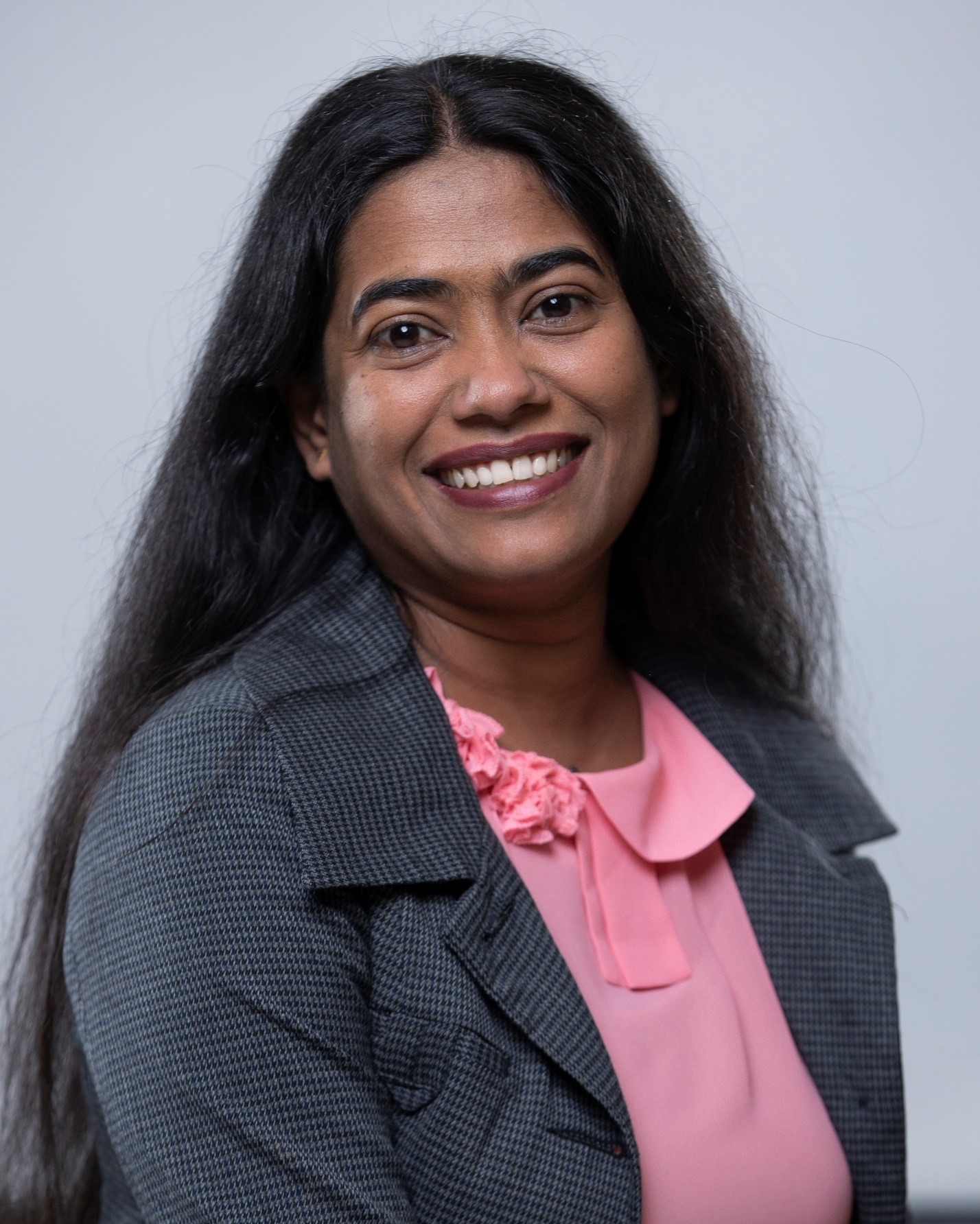 Professional headshot of a woman with long dark hair, wearing a grey blazer and pink blouse, smiling at the camera.