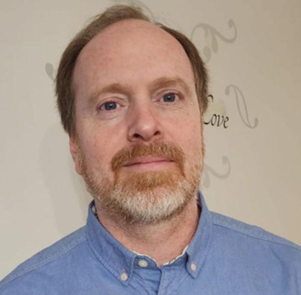 A close-up portrait of a man with a beard, wearing a blue button-up shirt, against a light-colored wall with decorative text.